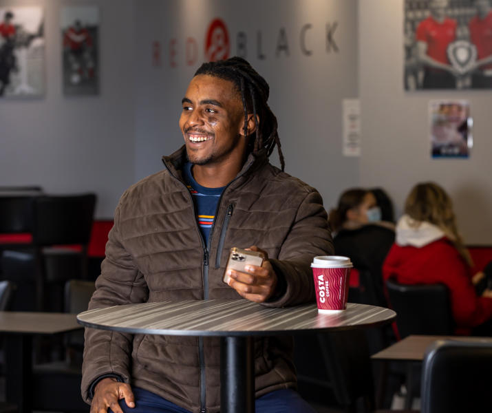 Image of international student standing at a table in a lounge on their phone with a hot drink